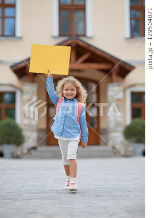 cheerful girl skipping toward school building holding handmade chart 129504071