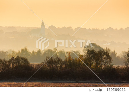 rural landscape on an autumn morning. row of trees along the empty fields. village with church on the hill behind the forest in the hazy distance rural landscape on an autumn morning. row of trees along the empty fields. village with church on the hill behind the forest in the hazy distance 129509021