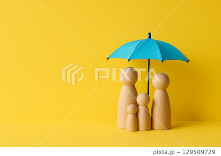 Wooden family figurines positioned under a small blue umbrella on yellow background representing safety and protection 129509729