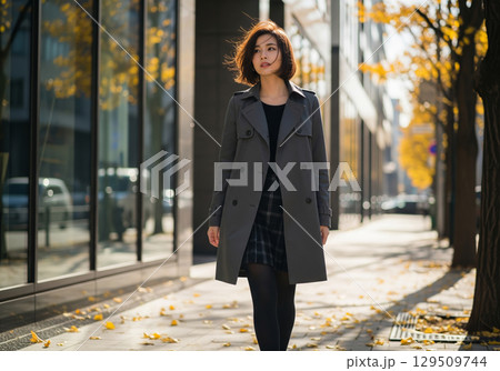 秋コーデ：都会の黄葉並木道を歩く、グレーのトレンチコートを着た風になびくショートヘアの女性モデル 129509744