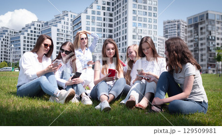A lively group of young women sits on the grass, dressed casually in light clothing, engaged with their smartphones under a clear blue sky, surrounded by urban architecture. 129509981