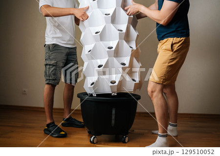 Cropped shot of two urban farmers placing main body of vertical hydroponic tower garden onto its base, working together to set up indoor space for cultivating fresh plants. Concept of home farming. 129510052