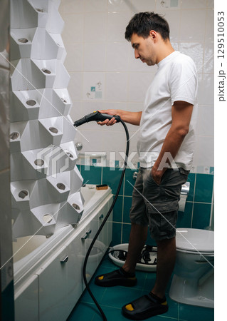 Vertical side view of man using steam cleaner to sanitize vertical hydroponic tower garden in bathroom, demonstrating modern and sustainable approach to indoor gardening. Concept of home gardening. 129510053