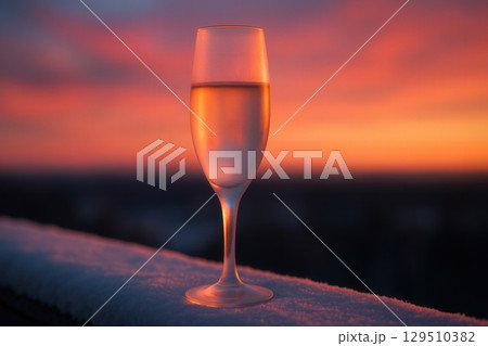 A champagne flute with bubbles on a snowy surface against a vibrant sunset sky 129510382