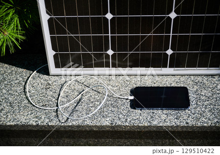 Solar panel set up on stone surface next to smartphone connected via charging cable. Practicality of using solar energy to power devices, demonstrating simple and sustainable charging solution. Solar panel set up on stone surface next to smartphone connected via charging cable. Practicality of using solar energy to power devices, demonstrating simple and sustainable charging solution. 129510422