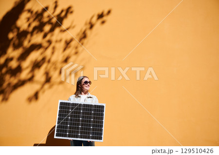 Woman holding solar panel for charging, stands against vibrant orange wall. Smiling girl wears sunglasses, white top. Artistic shadows cast by nearby trees branches on wall. 129510426