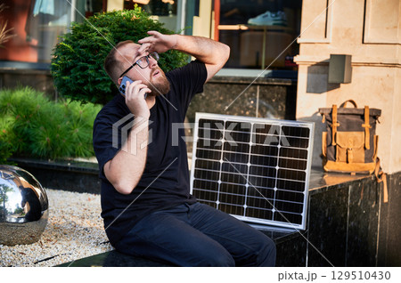 Bearded man talks on smartphone next to photovoltaic solar panel. Integration of sustainable renewable energy into everyday life, demonstrating practical use of solar power for charging devices. Bearded man talks on smartphone next to photovoltaic solar panel. Integration of sustainable renewable energy into everyday life, demonstrating practical use of solar power for charging devices. 129510430