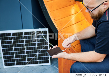 Cropped view of man connecting smartphone to photovoltaic solar panel. Integration of sustainable renewable energy into everyday life, demonstrating practical use of solar power for charging devices. Cropped view of man connecting smartphone to photovoltaic solar panel. Integration of sustainable renewable energy into everyday life, demonstrating practical use of solar power for charging devices. 129510431