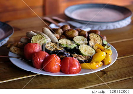 Grilled vegetables and sausages served on a white plate in a rustic dining setting during a summer gathering 129512003