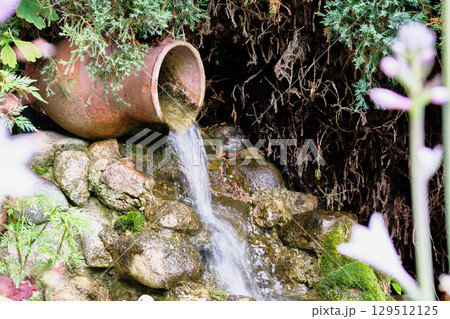 Water flows gently from a clay pot into a serene garden stream surrounded by lush greenery and vibrant plants Water flows gently from a clay pot into a serene garden stream surrounded by lush greenery and vibrant plants 129512125