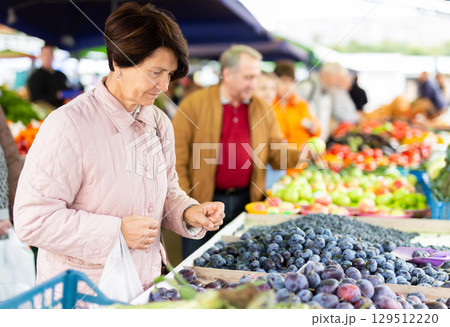 Positive mature woman in casual clothes picking fresh plums during shopping at grocery market 129512220