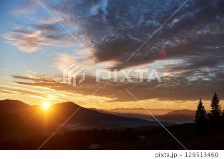 Sun rises behind hills, casting golden glow across sky filled with dramatic clouds. Valley below softly illuminated with mist, enhancing tranquil and picturesque landscape in the morning. Sun rises behind hills, casting golden glow across sky filled with dramatic clouds. Valley below softly illuminated with mist, enhancing tranquil and picturesque landscape in the morning. 129513460