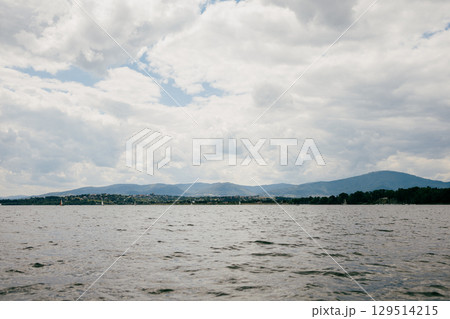 Lake reflecting cloudy sky with distant mountains and sailboats, offers serene view perfect for enjoying natura tranquility. Cloudy sky reflecting on lake, revealing distant mountains and sailboats Lake reflecting cloudy sky with distant mountains and sailboats, offers serene view perfect for enjoying natura tranquility. Cloudy sky reflecting on lake, revealing distant mountains and sailboats 129514215
