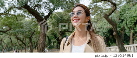 Young woman smiling while walking through a park on a sunny autumn day, showcasing a stylish trench coat and sunglasses 129514961