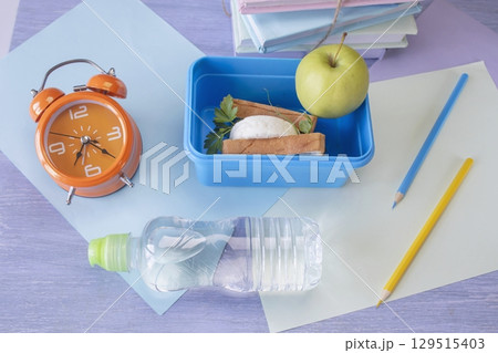 Back to school concept, school food. Lunch box with healthy sandwich, alarm clock, stack of books, apple and bottle of water on chalkboard background. Back to school concept, school food. Lunch box with healthy sandwich, alarm clock, stack of books, apple and bottle of water on chalkboard background. 129515403