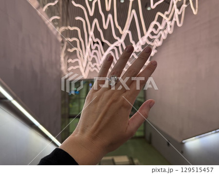 Landscape close up photo of a woman's hand wearing a large oval cut diamond engagement ring, held up against a modern escalator with artistic neon lights in the background. 129515457