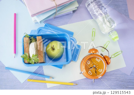 Back to school concept, school food. Lunch box with healthy sandwich, alarm clock, stack of books, apple and bottle of water on chalkboard background. Back to school concept, school food. Lunch box with healthy sandwich, alarm clock, stack of books, apple and bottle of water on chalkboard background. 129515543