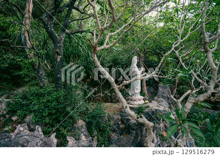 Serene marble statue of Guanyin in forest shrine with offerings at the base, Peaceful Buddhist female figure in jungle setting with altar and sacred atmosphere 129516279