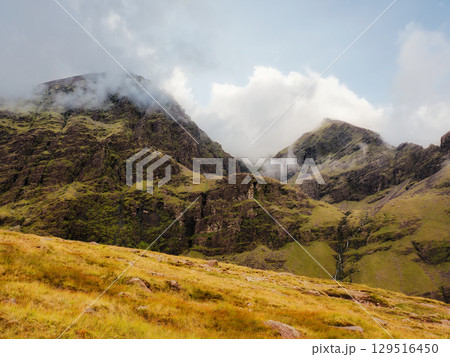 Rugged slopes rise steeply towards the summit of Carrauntoohill in County Kerry, Ireland. Low clouds swirl around the peaks, adding to the dramatic landscape. 129516450
