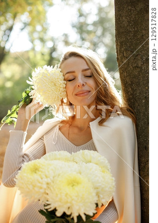 Portrait of curly young woman holding yellow flowers outdoors in autumn park. Woman enjoying warm autumn in a park. Woman wearing white coat. 129517828