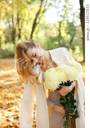 Portrait of curly young woman holding yellow flowers outdoors in autumn park. Woman enjoying warm autumn in a park. Woman wearing white coat. 129517833