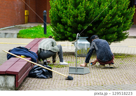 Two people cleaning weeds from pavement in public area with broom, dustpan, and trash bag Two people cleaning weeds from pavement in public area with broom, dustpan, and trash bag 129518223
