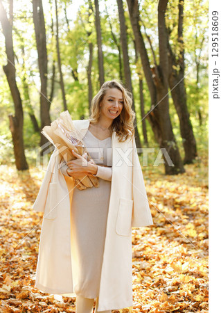 Portrait of curly young woman holding baguettes in craft paper outdoors in autumn. Woman enjoying warm autumn in a park. Woman wearing beige dress and white coat. Portrait of curly young woman holding baguettes in craft paper outdoors in autumn. Woman enjoying warm autumn in a park. Woman wearing beige dress and white coat. 129518609