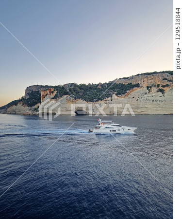 Vibrant evening seascape with a boat cruising away on the blue sea, near the Pirate Cave in Kemer at sunset. Beautiful Turkish mediterranean coastline lined with tree covered rocky cliffs at dusk 129518844