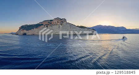 Vibrant evening seascape with a boat cruising away on the blue sea, near the Pirate Cave in Kemer at sunset. Beautiful Turkish mediterranean coastline lined with tree covered rocky cliffs at dusk 129518845