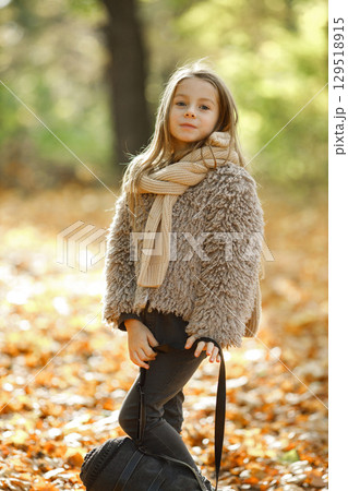 Portrait of little girl posing for a photo in autumn. Girl holding a black bag in her hand. Girl wearing grey sweater. Portrait of little girl posing for a photo in autumn. Girl holding a black bag in her hand. Girl wearing grey sweater. 129518915