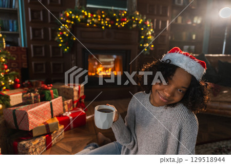 Merry Christmas. African American woman in Santa hat with cup of hot drink coffee tea near Christmas tree in dark interior. Girl in living room with Christmas tree and fireplace. Christmas eve at home Merry Christmas. African American woman in Santa hat with cup of hot drink coffee tea near Christmas tree in dark interior. Girl in living room with Christmas tree and fireplace. Christmas eve at home 129518944