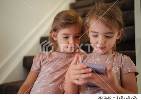 Two preschool age girls using a smartphone sitting on stairs at home 129519626