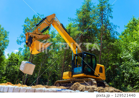 Excavator lifting large blocks stone at construction site surrounded by trees on works day Excavator lifting large blocks stone at construction site surrounded by trees on works day 129521440