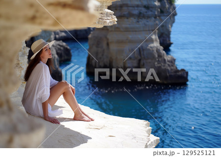 Relaxing by the Coastal Cliffs, a Woman Enjoys Tranquility Near the Turquoise Waters on a Sunny Day 129525121