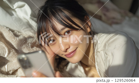 ベッドにうつ伏せになってスマホを見る女性 129526064