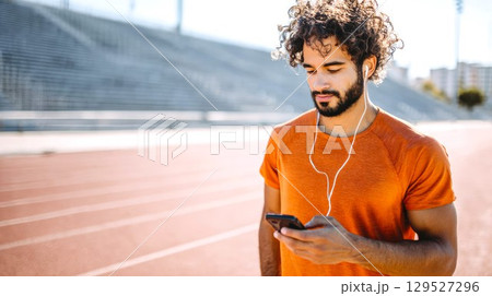 Young runner using phone and headphones at stadium. Curly sportsman listening to music during workout, morning run outdoors, sports and healthy lifestyle concept 129527296