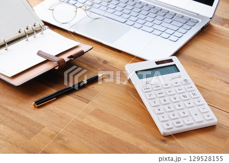 ノートパソコンと手帳、電卓を机に広げ、デスクワークをしているビジネスのイメージ 129528155