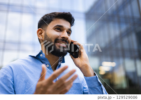 A smiling Indian man in a blue shirt talks on his cell phone with a modern building in the background. 129529086