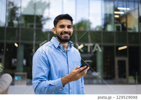 Smiling man in blue shirt holding a phone outdoors in front of a modern building. 129529098