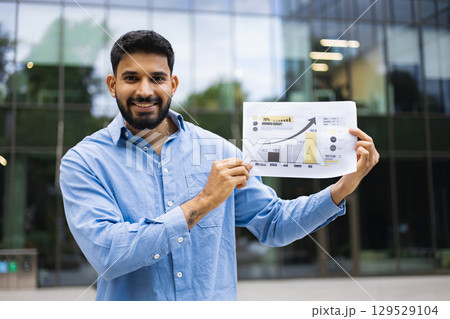 A smiling businessman presents a financial chart in front of a modern office building, symbolizing business growth and success. A smiling businessman presents a financial chart in front of a modern office building, symbolizing business growth and success. 129529104