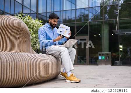 A businessman reviews financial data on a laptop and a printed chart outdoors in front of a modern building. A businessman reviews financial data on a laptop and a printed chart outdoors in front of a modern building. 129529111