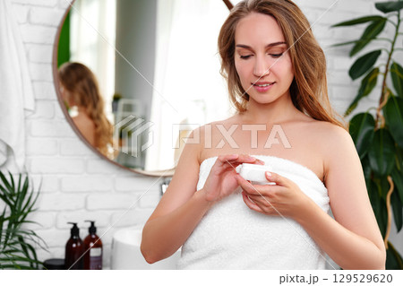 Woman applying skincare cream while wrapped in a towel in a bright bathroom in the afternoon 129529620