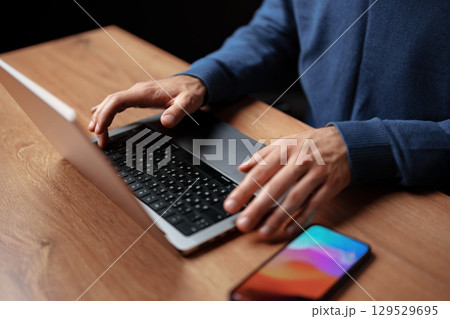 Hands typing on a laptop keyboard with a smartphone resting on a wooden desk during the evening Hands typing on a laptop keyboard with a smartphone resting on a wooden desk during the evening 129529695