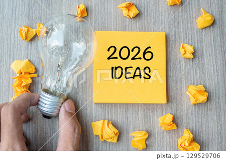 2026 Idea words on yellow note and crumbled paper with Businessman holding lightbulb on wooden table background. New Year New Start Creative, Innovation, Imagination, Resolution and Goal concept 129529706