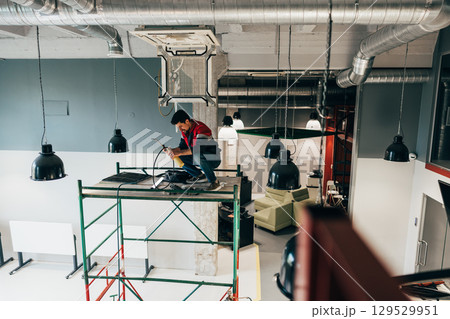 Worker performs electrical maintenance on scaffolding in modern office space during daylight hours 129529951