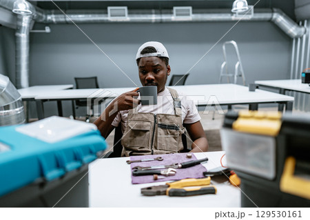 Worker enjoying a coffee break in a modern workshop with tools and equipment around during mid-morning hours 129530161