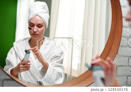 Woman applying skincare product while wearing a robe and towel in a bright bathroom setting Woman applying skincare product while wearing a robe and towel in a bright bathroom setting 129530178