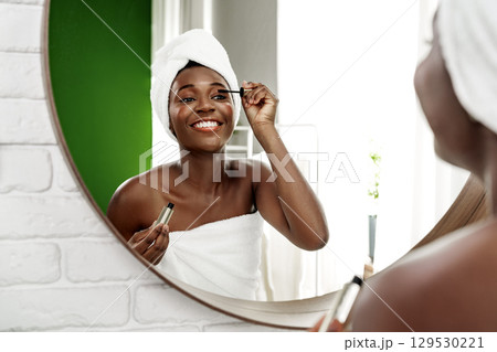 Woman applying mascara while smiling in a bright bathroom with green accents and a large mirror 129530221