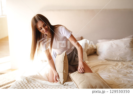 Woman arranging pillows on a bed in a sunlit bedroom during the morning hours 129530350