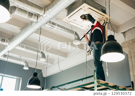 Technician performing maintenance on ceiling-mounted air conditioning unit in modern workspace 129530372